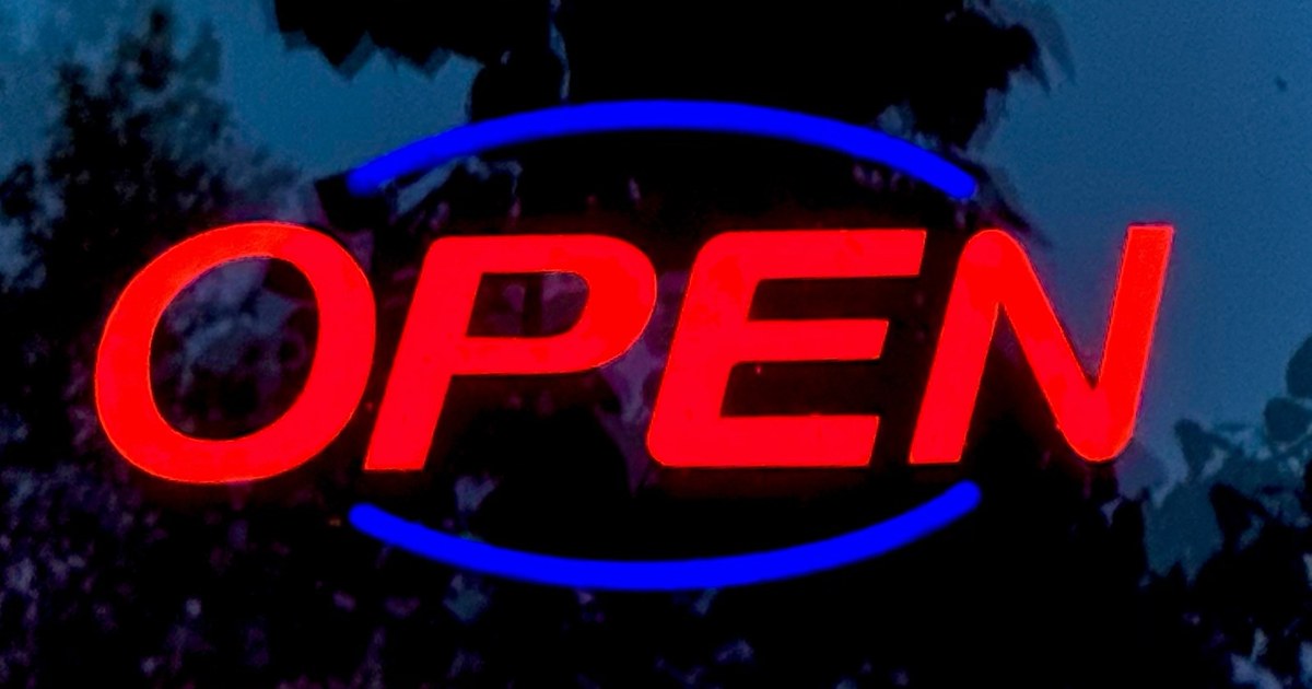 A glowing red and blue "OPEN" sign in a window at dusk, with dark trees reflected in the glass.